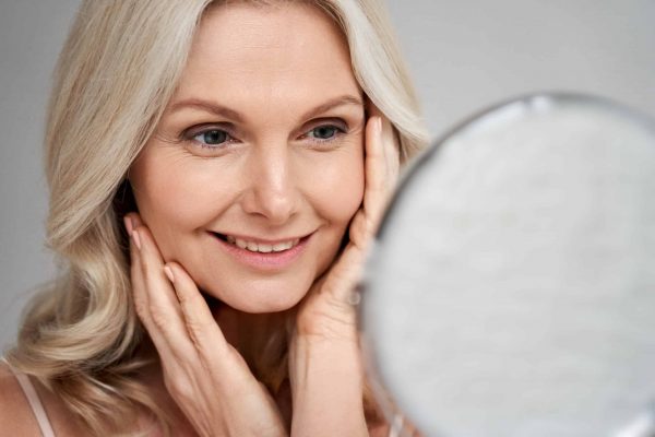 Happy 50s middle aged woman model touching face skin looking in mirror. Smiling mature older lady pampering, enjoying healthy skin care, aging beauty, skincare treatment cosmetic products concept.